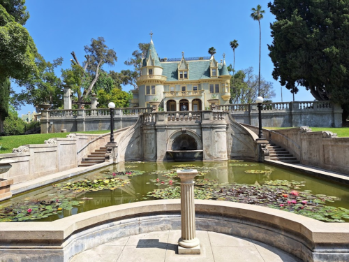 A grand mansion with a green roof, surrounded by lush trees and a pond filled with water lilies in the foreground.