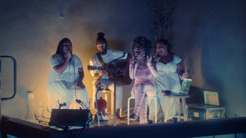 A group of four musicians perform together on a small stage, with a guitar and a warm, colorful backdrop.