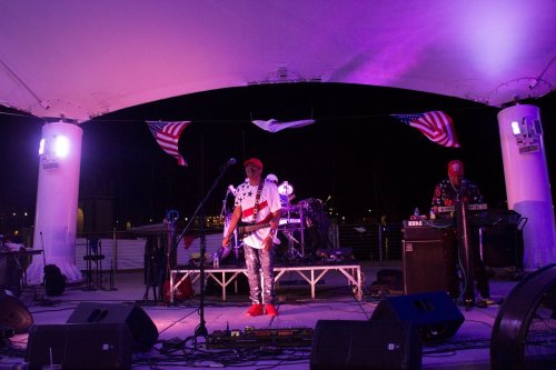 A band performs on stage at night, with American flags and colorful lights illuminating the scene.