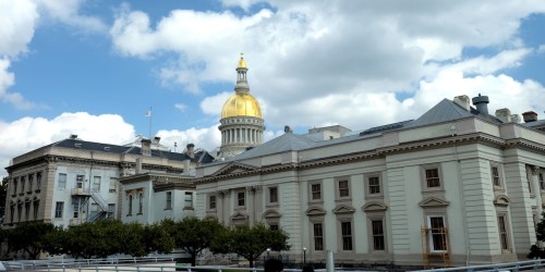 A historic building with a golden dome under a cloudy sky, surrounded by trees and a clear view of the architecture.