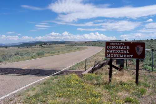 Sign for Dinosaur National Monument beside a road, with a clear blue sky and distant hills in the background.