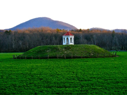 A small white gazebo with a red roof sits atop a grassy mound, surrounded by a green field and distant mountains.