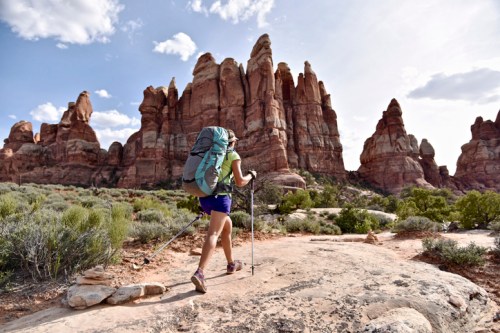 A hiker with a large backpack walks on a rocky trail, surrounded by towering red rock formations and green shrubs.