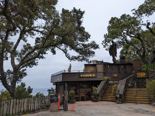 A rustic building named "Phoenix" surrounded by trees, with stairs leading up and a view of the ocean in the background.