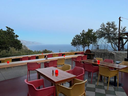 A scenic outdoor dining area with colorful chairs and tables overlooking a calm sea and distant mountains at dusk.