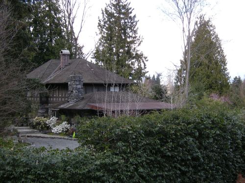 A stone and wood house surrounded by trees and shrubs, with a gravel driveway and a cloudy sky in the background.