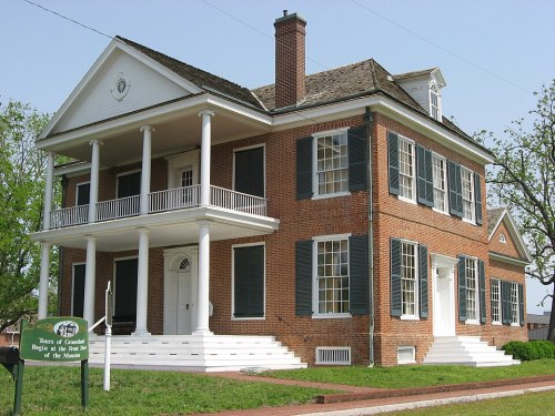 Historic brick house with white columns, green shutters, and a sign in front, surrounded by trees and a clear sky.