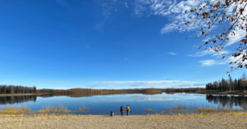 Three people stand by a calm lake under a clear blue sky, surrounded by autumn foliage and trees.
