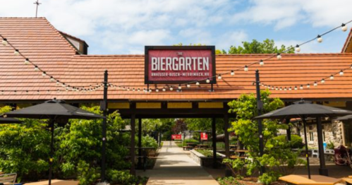 A sunny outdoor biergarten with a sign, string lights, and shaded seating areas surrounded by greenery.
