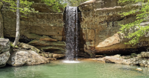 A serene waterfall cascading over rocks into a clear pool, surrounded by lush greenery and towering cliffs.