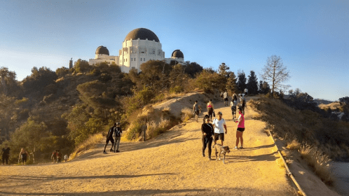 A scenic trail with people walking, leading to a large observatory on a hill, surrounded by trees and clear blue skies.