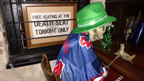 A skeleton in a green hat and sports jersey sits at a bar, next to a sign about free seating at the "Death Seat."