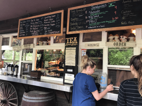 A rustic food stand with a menu board, featuring sandwiches and snacks, and two customers ordering at the counter.