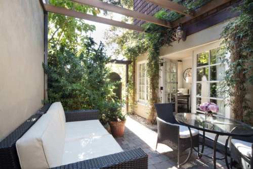 Cozy outdoor patio with a white sofa, glass table, and lush greenery surrounding a charming entrance.