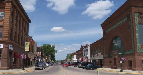 A sunny street view of a small town with shops, cars parked along the sides, and a blue sky with fluffy clouds.
