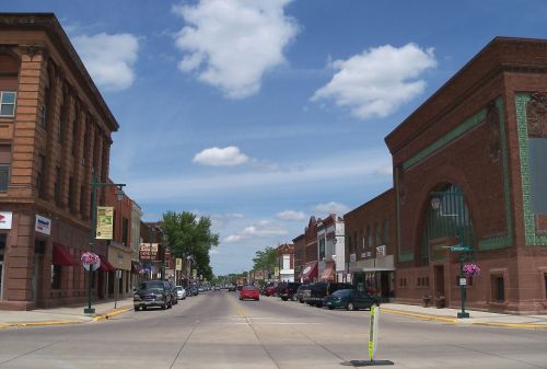 A sunny street view of a small town with shops, trees, and fluffy clouds in the blue sky.