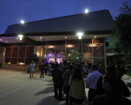 A line of people waiting outside a building at dusk, with lights illuminating the entrance.