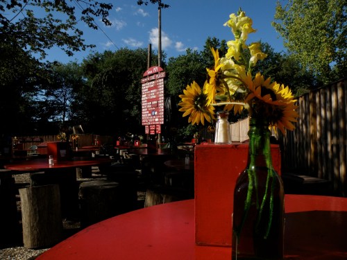 A sunny outdoor dining area with red tables, wooden seating, and a vase of sunflowers on a table.