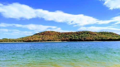 A serene landscape featuring a calm blue lake and lush green hills under a bright blue sky with scattered clouds.