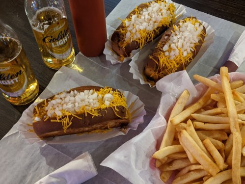 Three loaded hot dogs with onions and cheese, two beers, and a side of golden fries on a table.