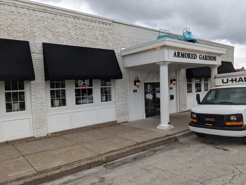 Exterior of Armored Gardens restaurant with a U-Haul truck parked outside, featuring white brick and black awnings.
