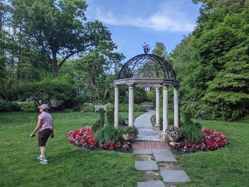 A person walks past a decorative gazebo surrounded by colorful flowers in a lush garden.