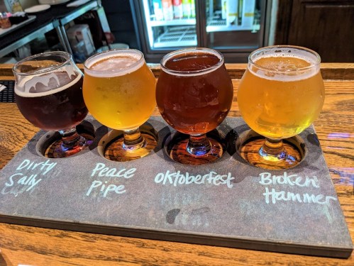 A flight of four craft beers in small glasses on a wooden tray, labeled with their names.