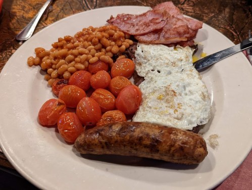 A plate of breakfast featuring baked beans, crispy bacon, fried eggs, cherry tomatoes, and a sausage.