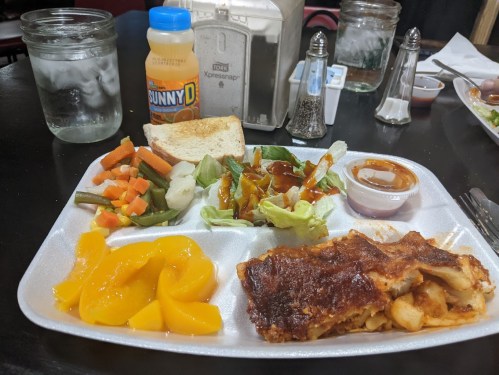 A tray with a slice of lasagna, salad, mixed vegetables, peaches, a slice of bread, and a drink on a table.
