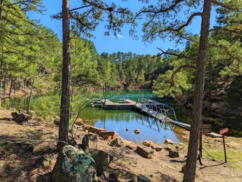 A serene lake surrounded by trees, featuring a wooden dock and rocky shoreline under a clear blue sky.