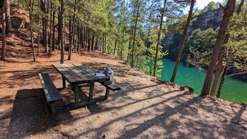 A picnic table in a wooded area beside a green lake, surrounded by tall trees and rocky cliffs.