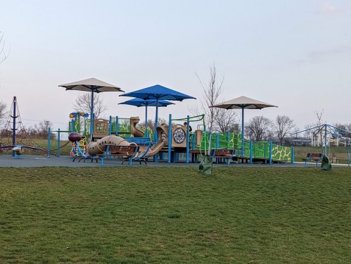 Colorful playground with slides and climbing structures, shaded by large umbrellas, set in a grassy area.