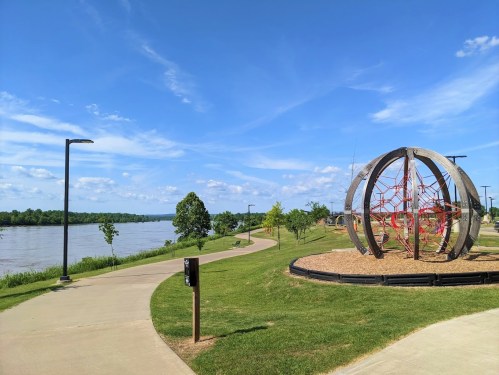 A park path curves alongside a river, featuring a large playground structure and green trees under a blue sky.