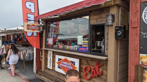 A wooden sandwich stand with an "Open" sign, surrounded by colorful merchandise and people walking by.