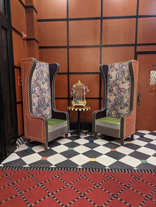 Two ornate chairs with colorful upholstery face each other beside a small table and a decorative birdcage, set on a patterned floor.