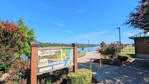 A park entrance sign with a view of a calm lake and trees under a clear blue sky. Pathway leads into the park.
