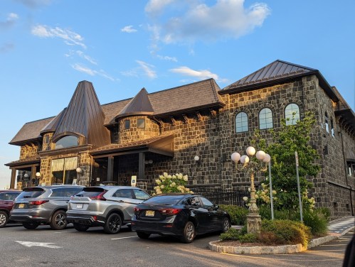 A large stone building with a peaked roof, surrounded by parked cars and greenery under a blue sky.