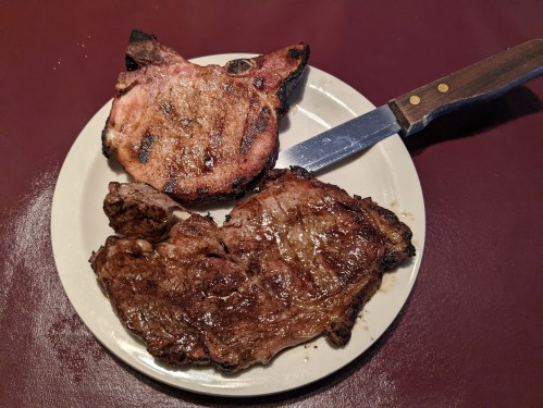 A plate with two grilled meat cuts: a pork chop shaped like a pig's head and a juicy beef steak, with a knife beside them.