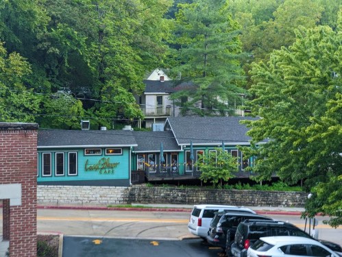 A cozy café nestled among trees, featuring a deck and surrounded by greenery, with parked cars in the foreground.