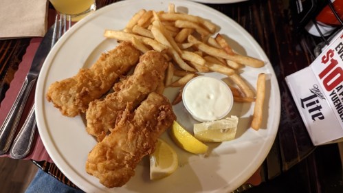 A plate with three fried fish fillets, French fries, lemon wedges, and a small cup of dipping sauce.