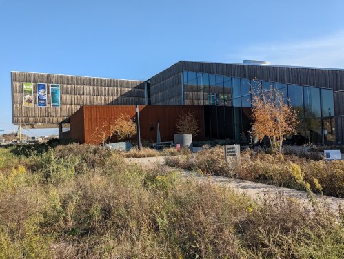 Modern building with wooden exterior and large glass windows, surrounded by landscaped greenery under a clear blue sky.