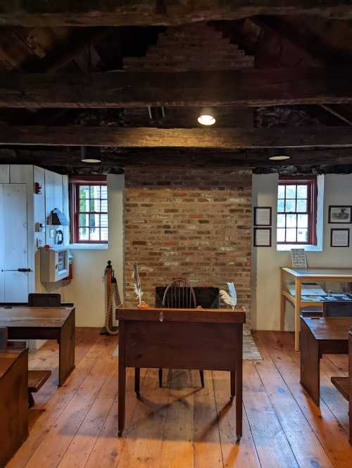 A rustic classroom with wooden desks, a chair, and a brick fireplace, featuring exposed beams and windows.