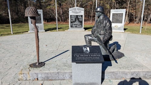 A soldier statue at a veterans memorial park, with plaques and a helmet nearby, honoring T. Sgt. Frank E. Barone.