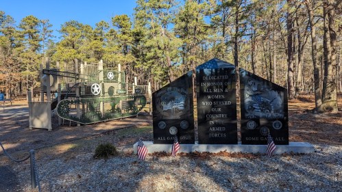 Memorial honoring military service members, featuring a tank sculpture and engraved stone markers, surrounded by trees.