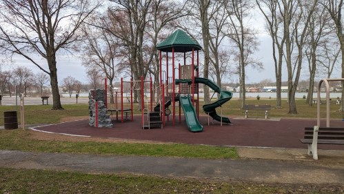A playground with a slide, climbing structure, and swings, surrounded by trees and open grass.