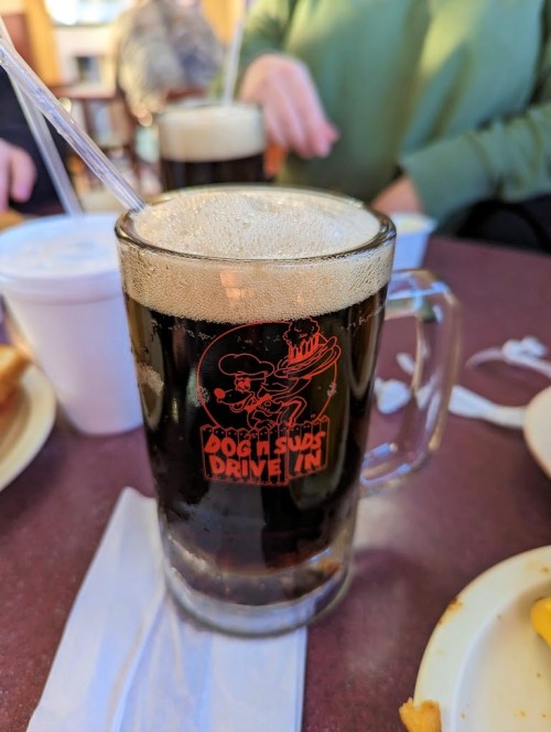 A frothy dark beer in a glass mug with a logo, placed on a table with food and other diners in the background.