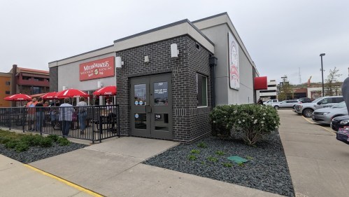Exterior view of a restaurant with outdoor seating, umbrellas, and a parking lot on a cloudy day.