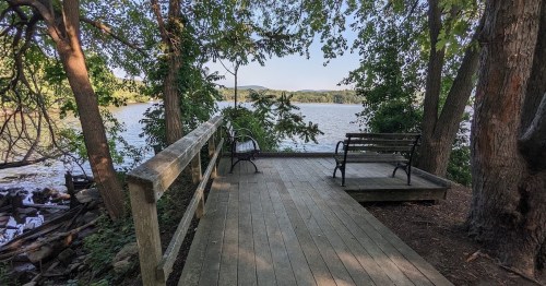 A wooden deck by a river, surrounded by trees, with two benches facing the water and distant hills in the background.