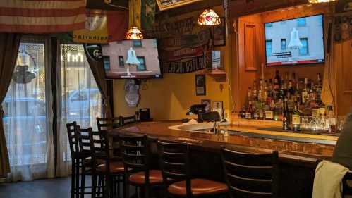 A cozy bar interior with a wooden counter, empty chairs, and two TVs displaying news, surrounded by sports memorabilia.