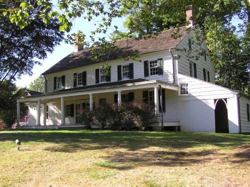 A two-story white house with green shutters, a porch, and a large lawn surrounded by trees.